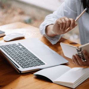 Individual holding a pencil and notebook while sitting at a desk with a laptop and mouse.