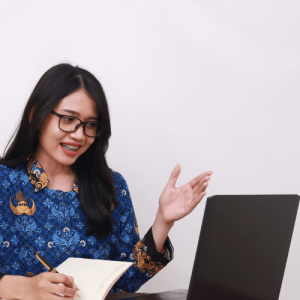 Woman in a blue patterned blouse speaking while using a laptop and taking notes in a notebook.